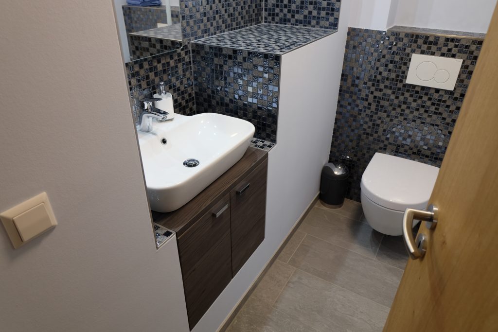Vorteilhaft genutzter Platz einer kleinen Gäste-WC mit dunklen Mosaikelementen, einer Toilette und einem Waschbecken mit Unterschrank.