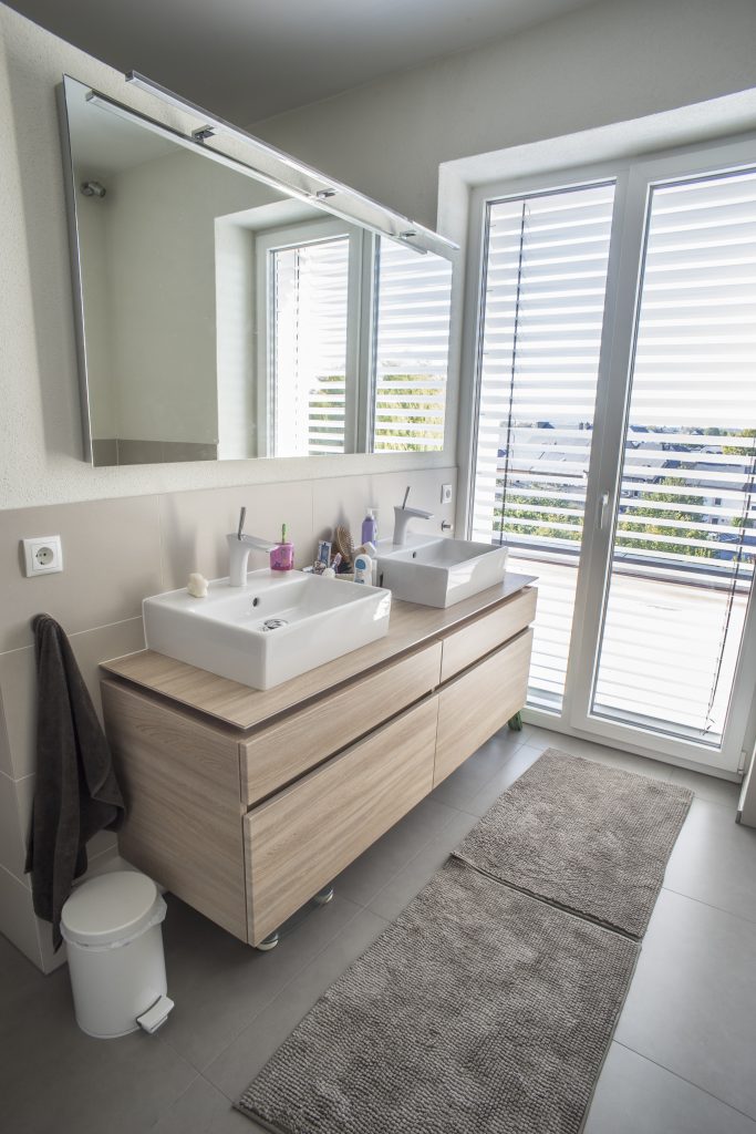 Modernes Bad mit einem Waschtisch, auf dem sich zwei eckige Waschbecken befinden, daneben ein bodentiefes Fenster, was auch als Tür in den Außenbereich dient.