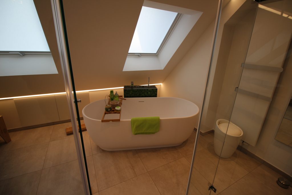 Freistehende Badewanne unter einem Dachfenster mit indirektem Licht an der Wand dahinter.