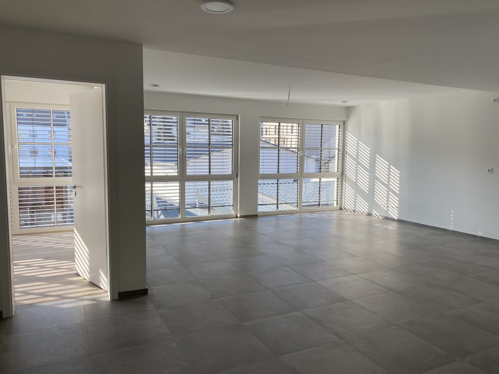 Unmöbliertes großes und offenes Wohnzimmer mit vier bodentiefen Fenstern, weißen weißen Wänden und grauen Bodenfliesen.