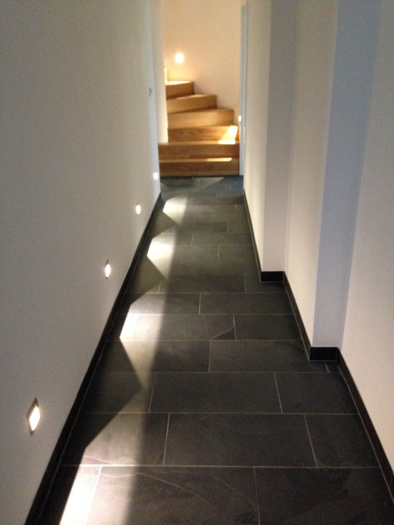 Weißer Hausflur mit eingebauten Lichtspots an den Wänden, grauen Bodenfliesen und einer Holztreppe am Ende des Gangs.