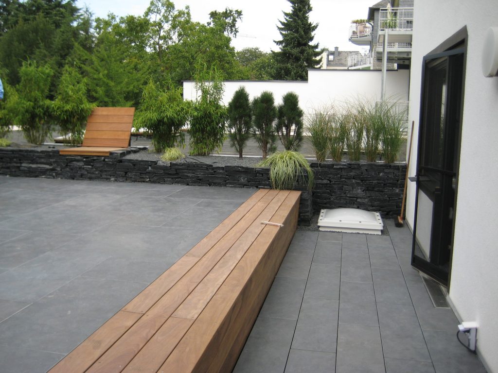 Moderne Terrasse mit grauen Bodenfliesen und einer Schiefermauer, auf der sich eine hölzerne Sitzgelegenheit mit Rückenlehne befindet.