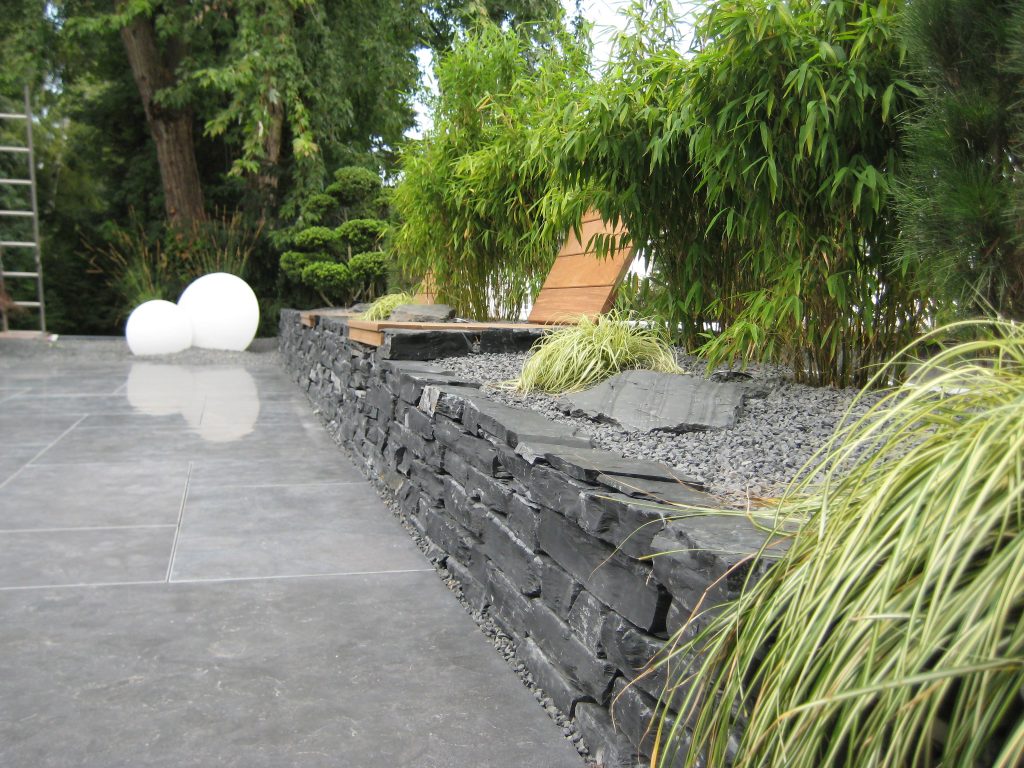 Graue Terrasse mit weißen Beleuchtungselementen und einer Schiefermauer, auf der sich eine hölzerne Sitzmöglichkeit mit Rückenlehne befindet, dessen Bereich mit vielen grünen Sträuchern dekoriert ist.