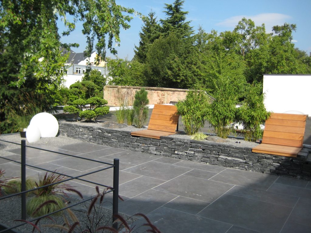 Graue Terrasse mit weißen Beleuchtungselementen und einer Schiefermauer, auf der sich eine hölzerne Sitzmöglichkeit mit Rückenlehne befindet, dessen Bereich mit vielen grünen Sträuchern dekoriert ist.