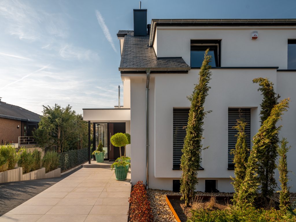 Frontansicht eines sanierten Hauses im modernen Baustil mit Pinienbäumen im Vorgarten und einer Auffahrt links am Haus entlang, die nach unten führt.