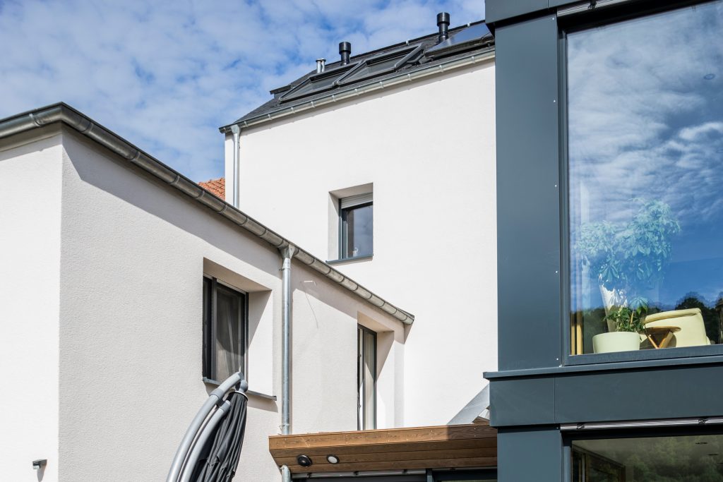 Haus mit weißer Fassade und grauem Blechdach, auf dem sich zwei Dachfenster und eine Photovoltaikanlage befinden.