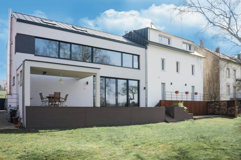 Außenansicht eines Anbaus mit einer durchgehenden Fensterfront im Obergeschoss und einer Terrasse auf einer Empore vom Garten aus.