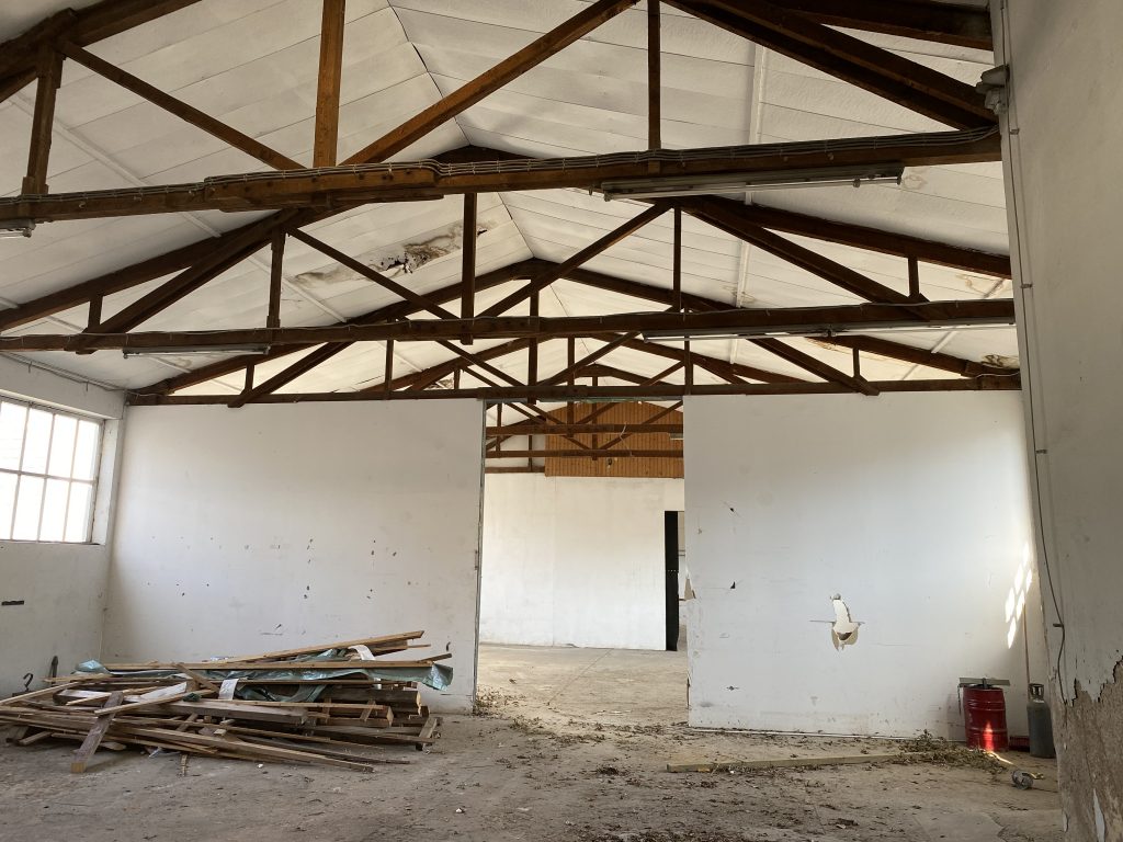 Alte heruntergekommene Lagerhalle, in der Balken an der Decke sind und Schutt auf dem Boden liegt.