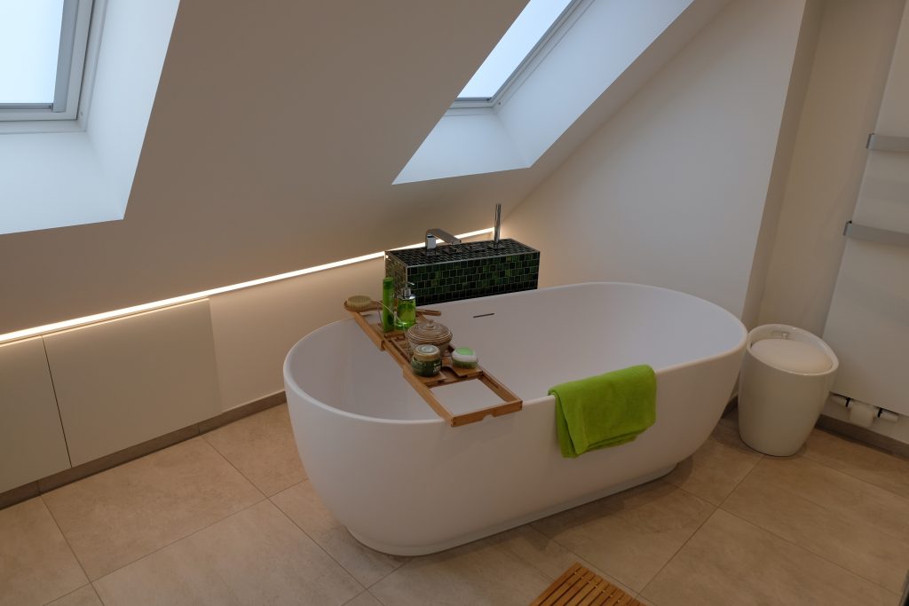 Eine ovale freistehende Badewanne ist unter zwei Dachfenstern auf sandfarbenem Boden platziert.