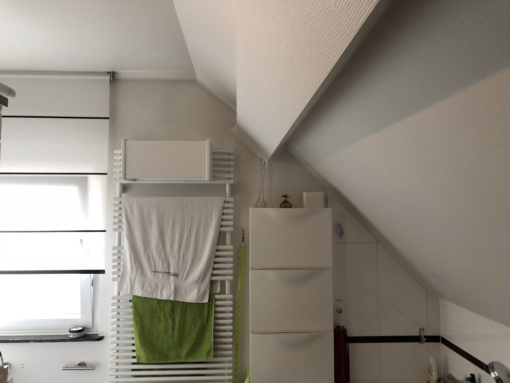 Weißes Dachgeschossbadezimmer mit einem Fenster an der linken Seite,, einem weißen Sprossenheizkörper in der Mitte, auf dem Handtücher platziert sind und einem weißen Wandschrank rechts.